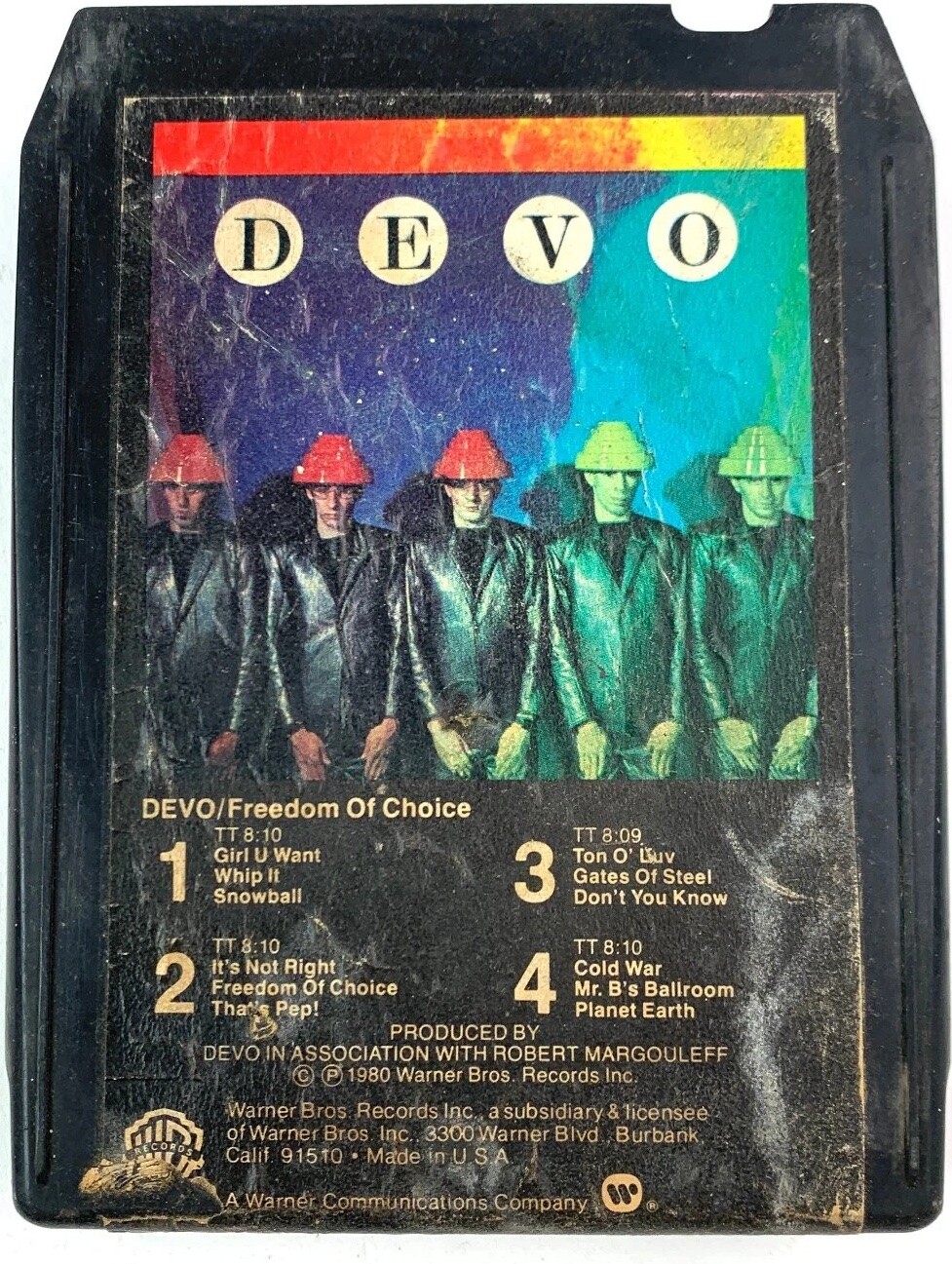 A photo of a vintage black 8-track cartridge tape entitled Freedom Of Choice by DEVO. Inset photo above the track listing & credits shows the band members standing stiffly with arms down in a line against a dark blue wall. Each band member is clad in a black leather jacket and Energy Dome™. Light water damage has faded the colours on the right side of the front label, resulting in a pretty rainbow effect to the original red/blue/black colour scheme.